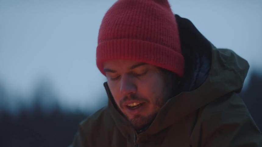 Man with a red beanie and a green coat stands next to the campfire on a cold cloudy day