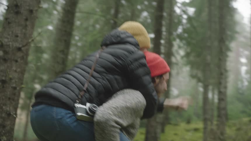 Man with a red beanie and a grey sweater carries a woman with a yellow beanie and black coat on his back in the forest