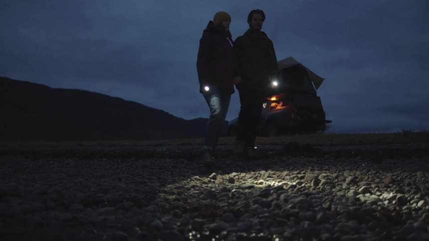 A couple holding flashlights walking outside at night near a truck with camping gear