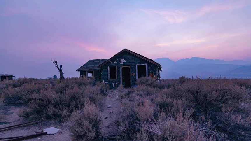 The exterior of an abandonded house in the middle of nowhere with low sun in a pink and blue sky with hills in the background