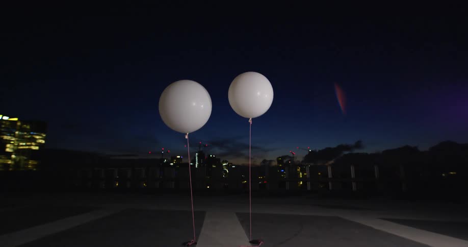 Two white balloons outside at night in front of a distant city view