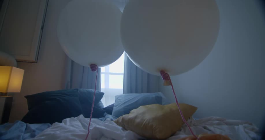 Two helium filled white balloons float over a bed with rumpled sheets
