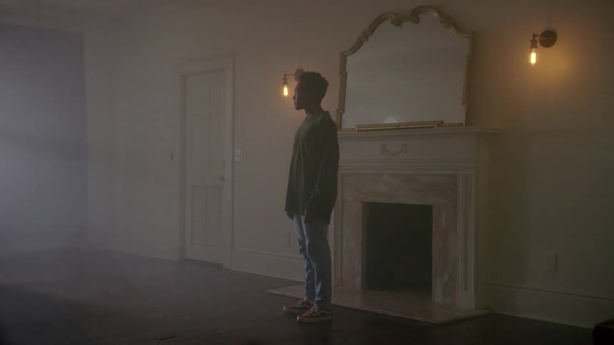 Young man standing in an empty living room in front of a fireplace with a mirror above it looking towards a window