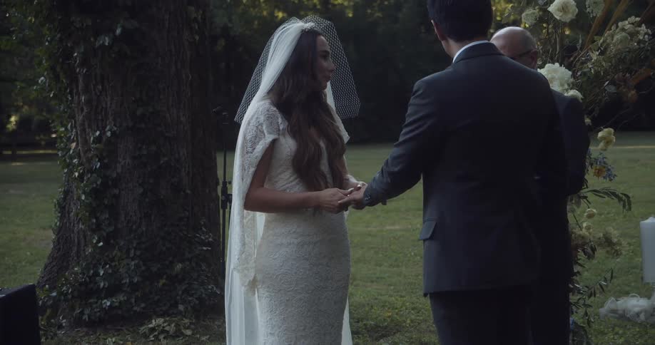Bride and groom standing at an outdoor alter at daytime putting wedding bands on each others fingers next to a priest