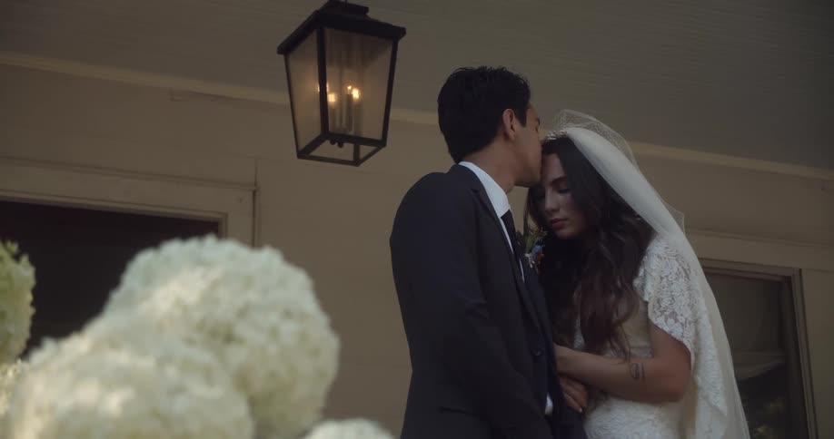 A newly married couples stands close as the husband kisses the wife on her forehead