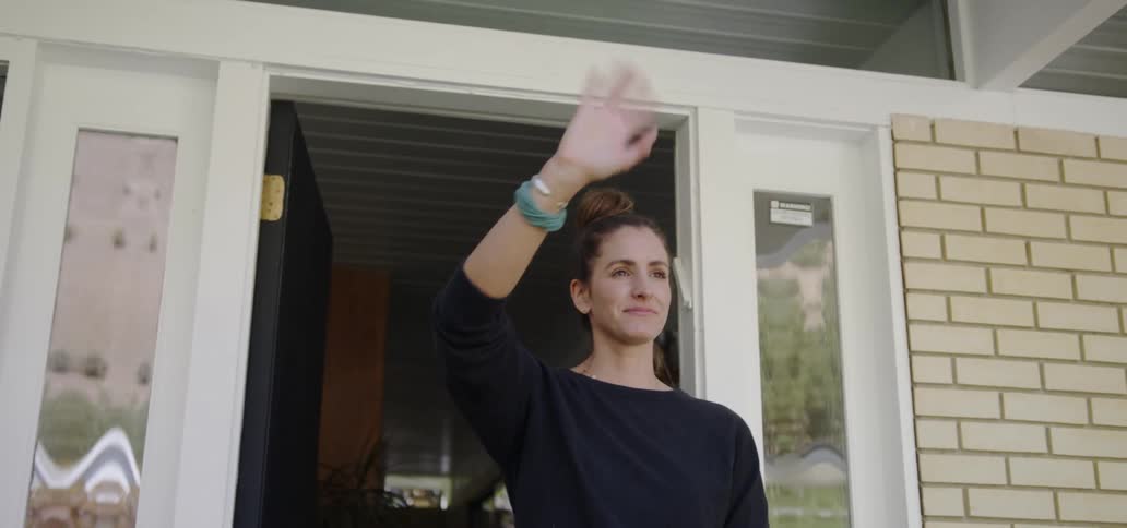 A woman waves good bye as she stands outside a brick house at the front door