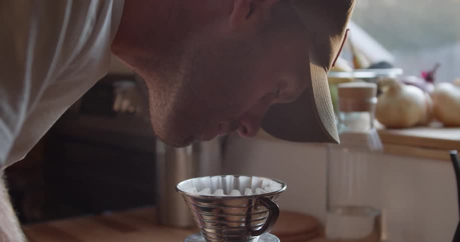 Male wearing a brown hat and white shirt bending over a coffee maker having a smell of the coffee