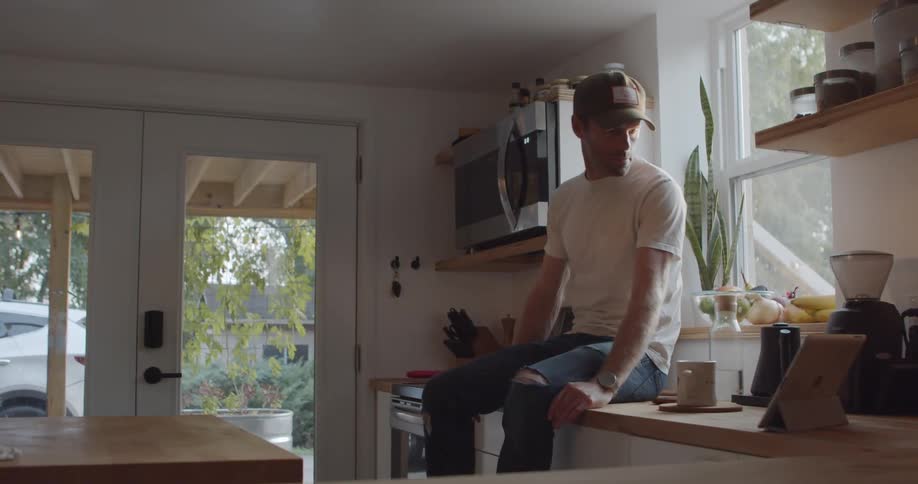 Man with a cap a white shirt and ripped jeans sits on kitchen counter and takes a tablet to use in the kitchen