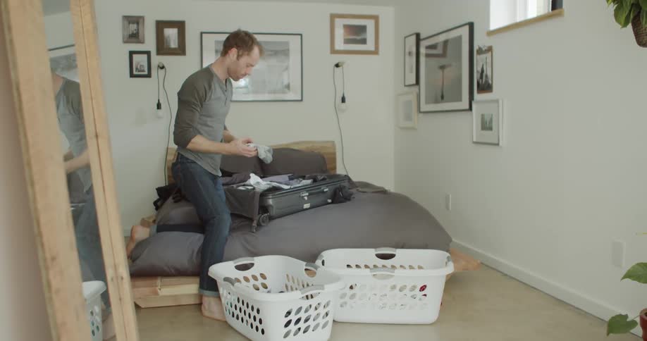 Man half kneels on bed putting clothes from laundry basket into open suitcase