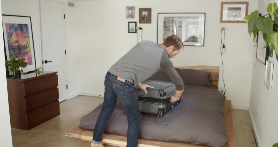 A barefoot man lifts a travel suitcase onto a bed and before unzipping and opening it