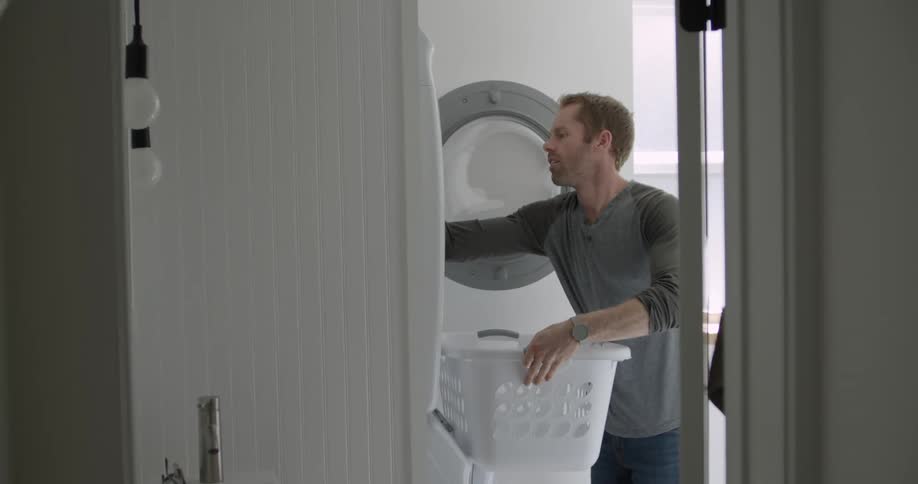 Man walks into a laundry room in his house carrying a laundry basket and then takes clothes out of a dryer