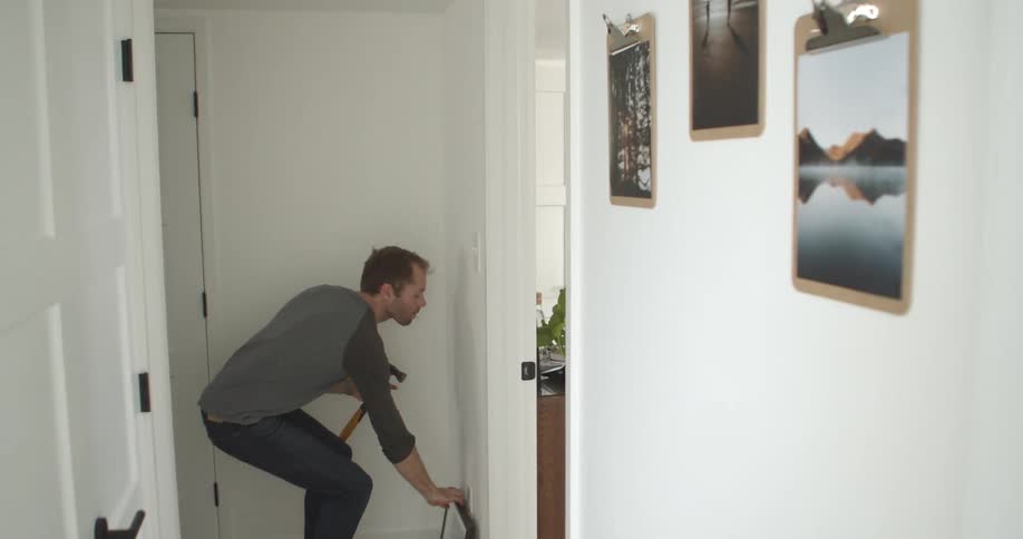 Man stands in hallway determining placement of framed picture then hammering in nail