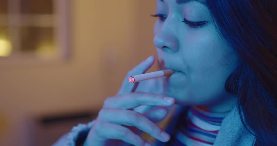 Woman with dark wavy hair and a denim jacket smokes a cigarette