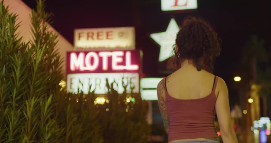 Woman with tattoos on her arm walking down a street at night next to a motel
