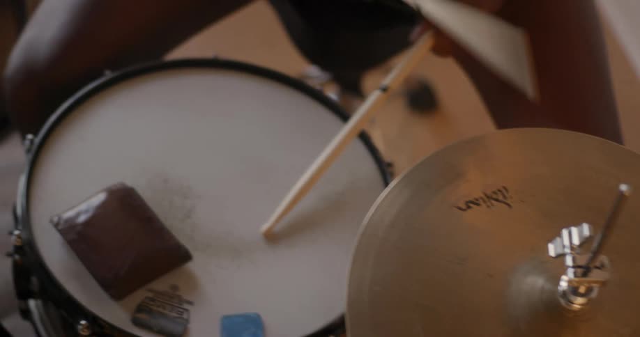 A person in shorts sits on a stool while playing a drum set with wooden drum sticks