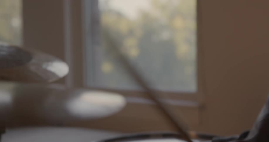 A man plays the drums in a room with sunlight coming through the windows
