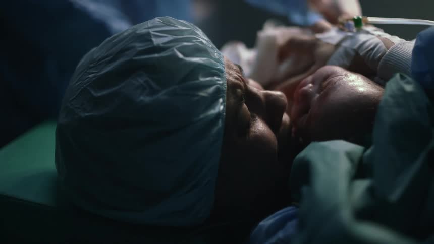 A woman holds her newborn in her arms as she lies on a bed in an operating room as she sobs