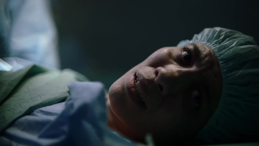A woman lies on a hospital gurney in sterile garb peering to her left with a fearful expression