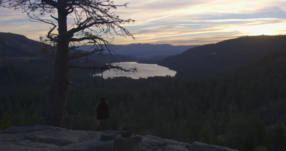A woman sits on a swing outside above a forest and lake