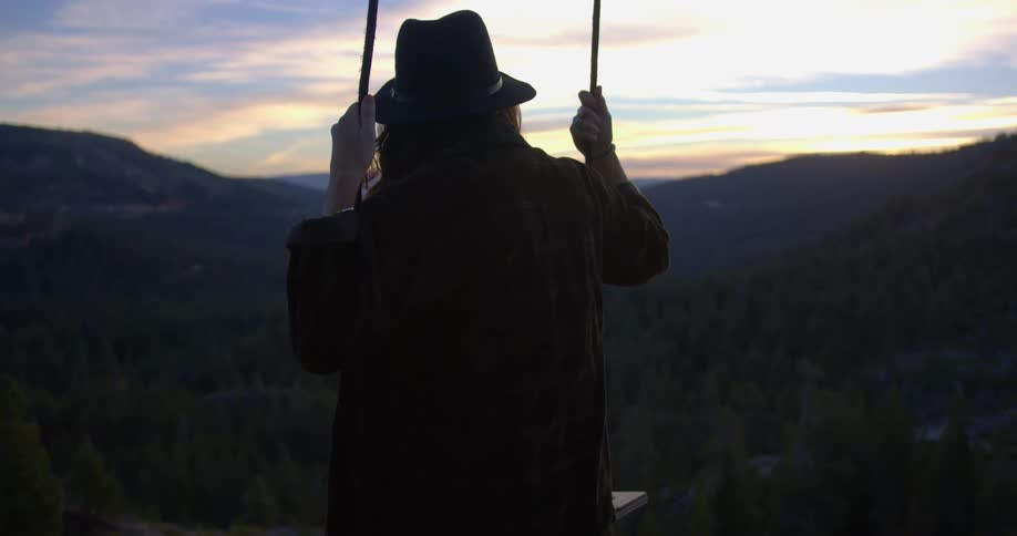 A woman swings on a swing overlooking mountains and water at sunset
