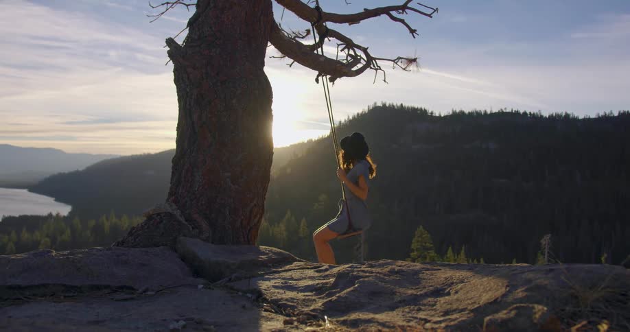 Woman swinging from a tree swing by a cliffside next to a sunset