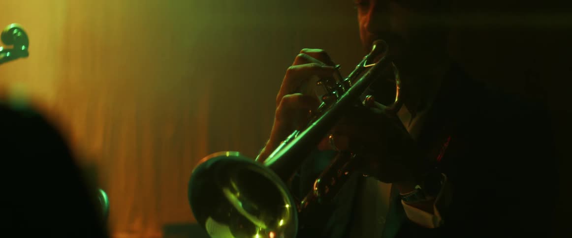 Three men wearing tuxedos play musical instruments including a trumpet and cello on stage