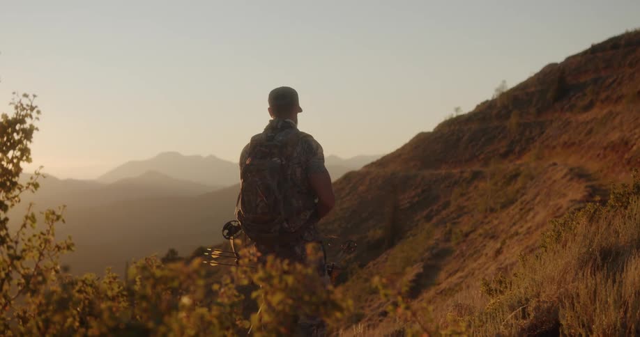 A hunter lifts up his bow and arrow while standing on the side of a hill
