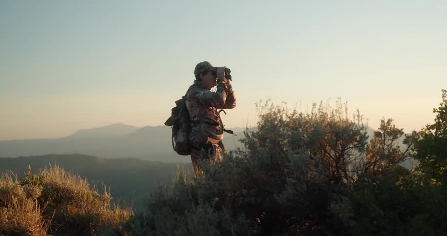 Man looks into the distance with binoculars standing near bushes in grassy area with mountain range in the background at sunset