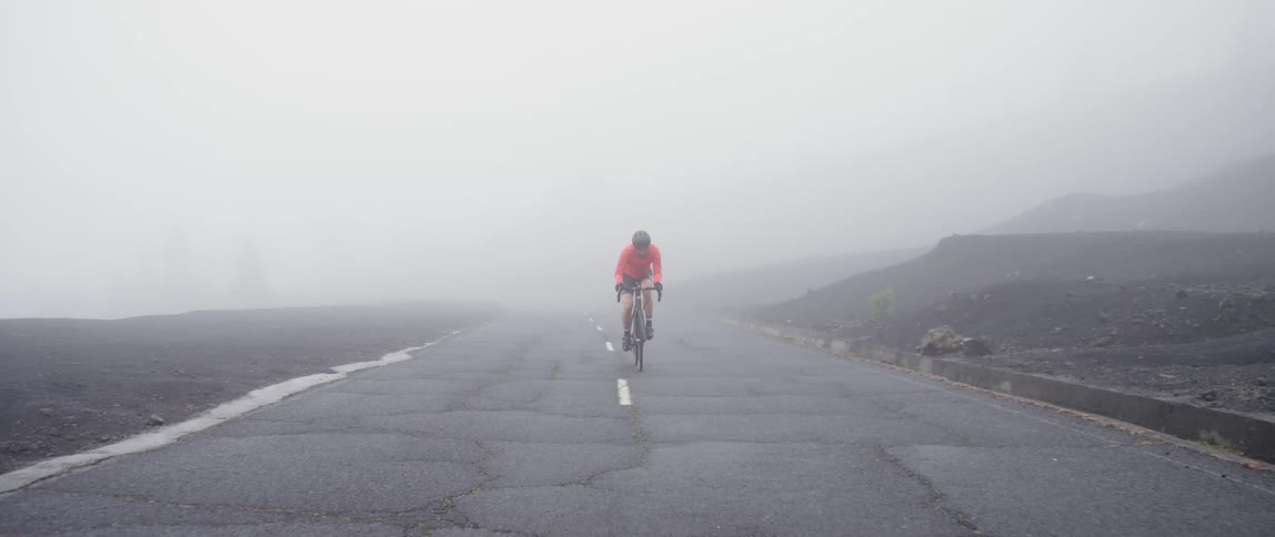 A person rides a bicycle up hill through heavy fog on a quiet rural road