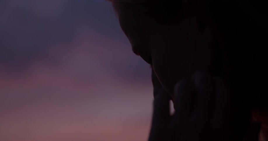 A young woman looks down at the ground and puts her head in her hands as the sun sets in front of a city skyline in the distance