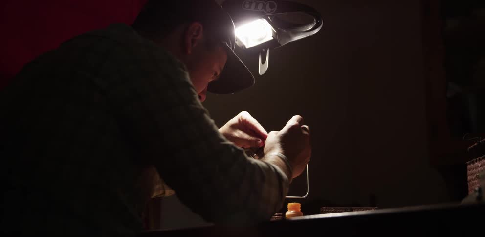 Man in a dimly lit room crafting a fishing bait under a single lamp