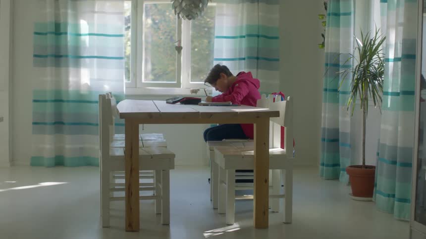 A boy works on homework on a dining table indoors during the day