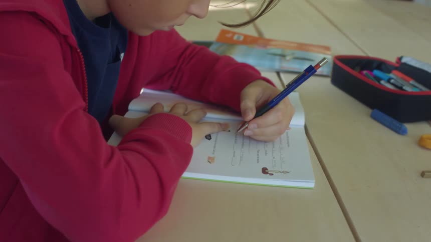 A boy sits at a table writing in a book with other school items around him