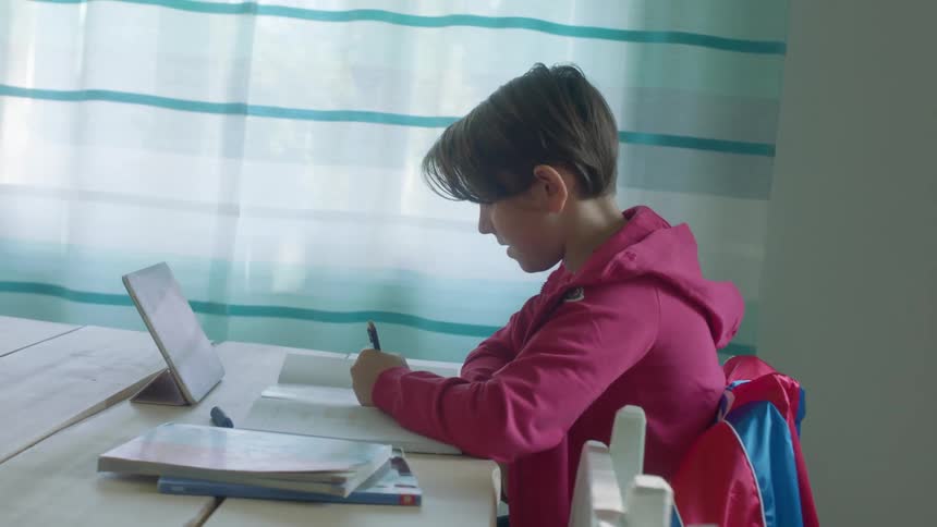 A child is sitting at the tablet video chatting on a tablet with homework in front of them