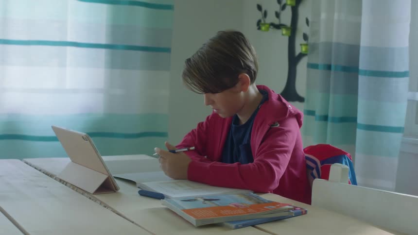 A young boy does school work at home while watching his teacher on a tablet