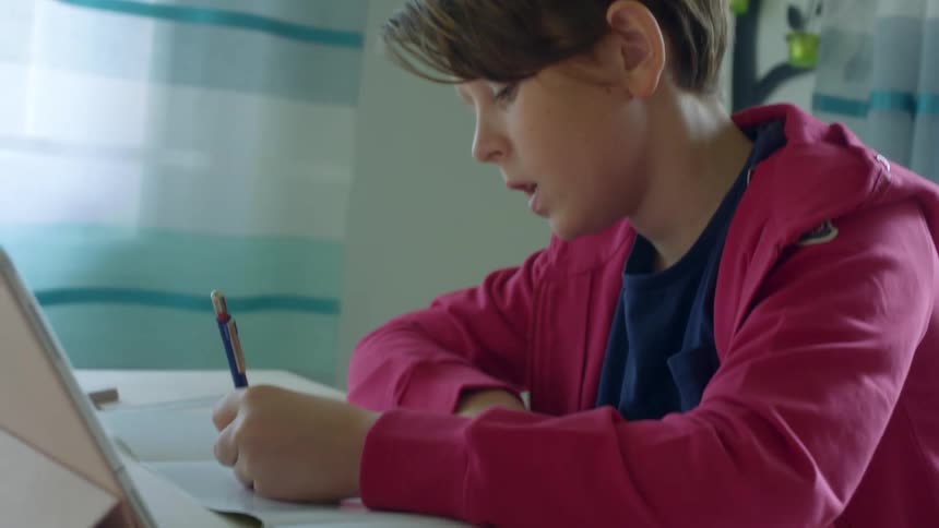 A boy sits at a table writing in a book in front of a tablet propped up