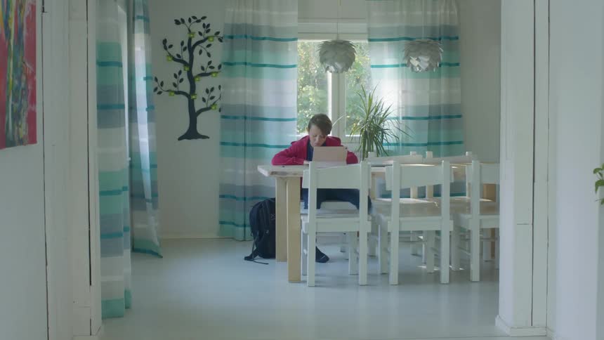 Boy sits at table doing homework on laptop in front of window with bookbag on the floor beside him