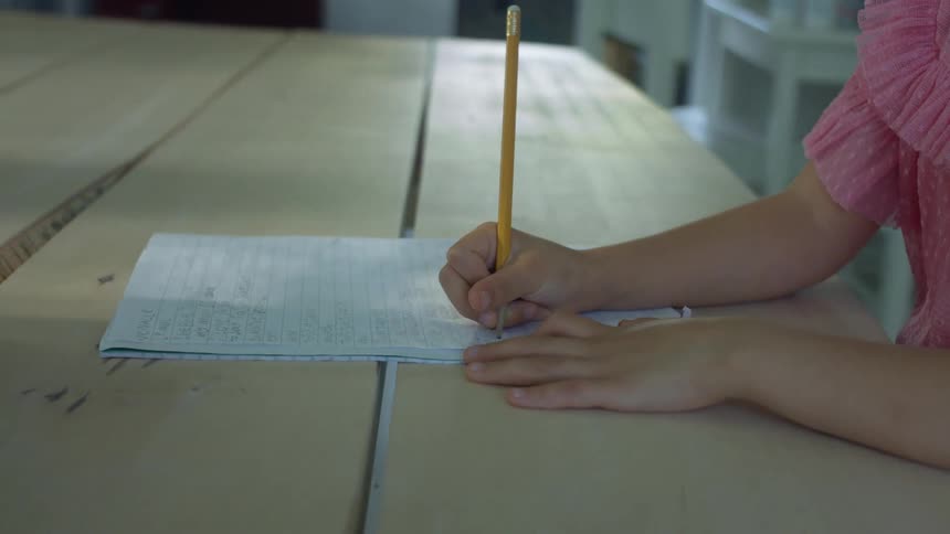 A girl sitting at a wooden table writes in a notebook with a pencil while participating in a video conference on a tablet on a stand