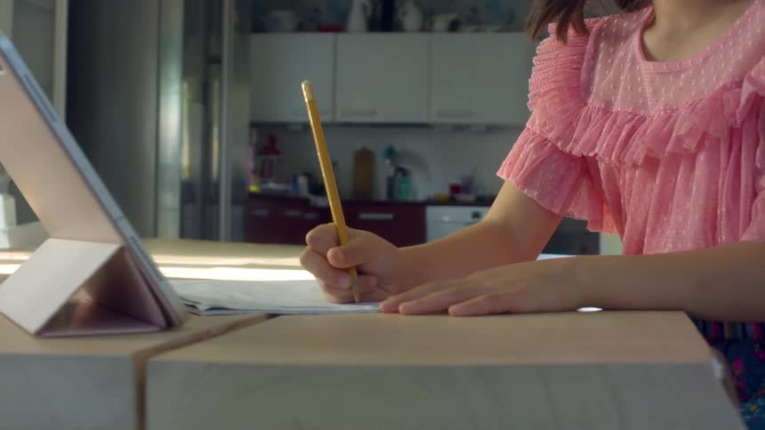 A young girl sits at a home table having a video call and writing on a piece of paper