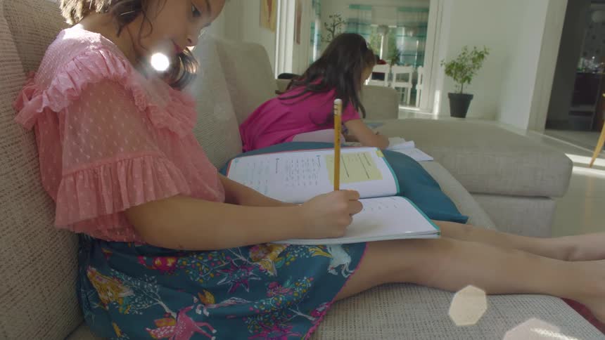 Two little girls sit on a couch at home and write in notebooks