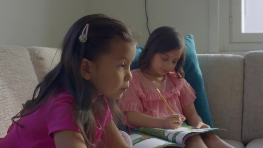 Two young girls sit on a couch at home and do school work