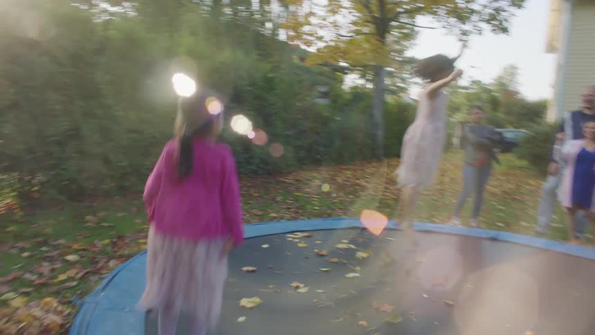 Three children jump on a trampoline in the backyard of a house as adults watch from nearby