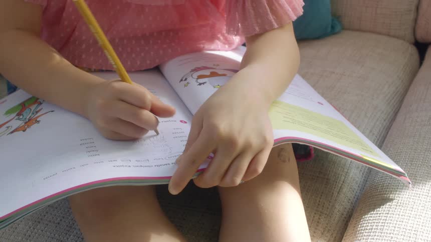 A young girl sits on her couch and fills in a worksheet with a pencil