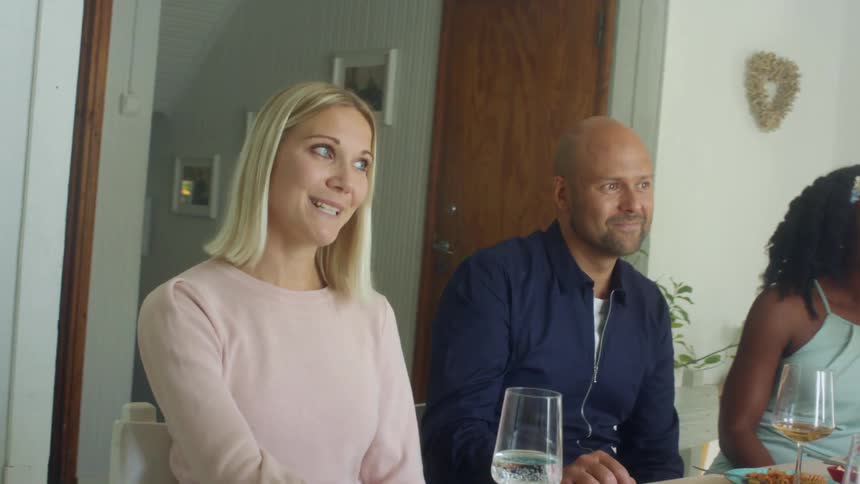 Two women and a man sit at a home dinner table talking to other people in the room