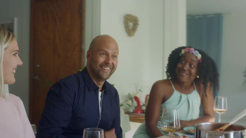 A group of people sit at a table in a home with food and wine and talk and laugh