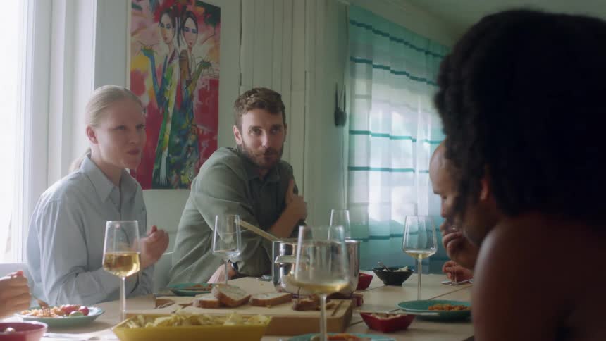 A group of men and women sit at a home dinner table having conversation
