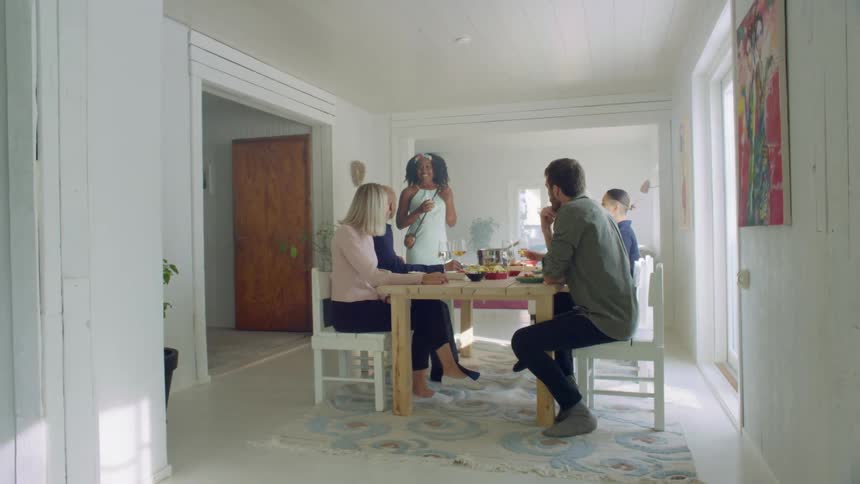 A woman stands up and gives a toast as a group of friends sit at a table in a home enjoying a meal and drinks