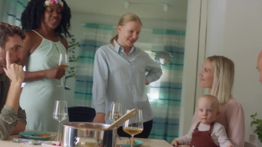 A group of men and women stand and sit around a dinner table one woman holding a baby