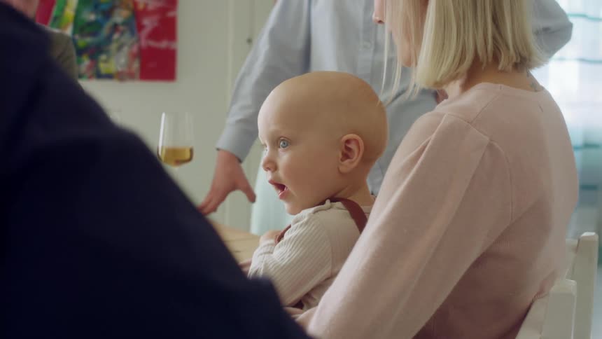 A blonde woman holds a baby while sitting at a dining table with three other people with one standing at the end of the table