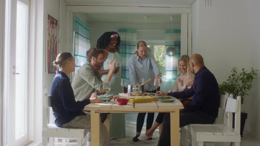 A group of men and woman gather around a home dinner table admiring a woman holding a baby